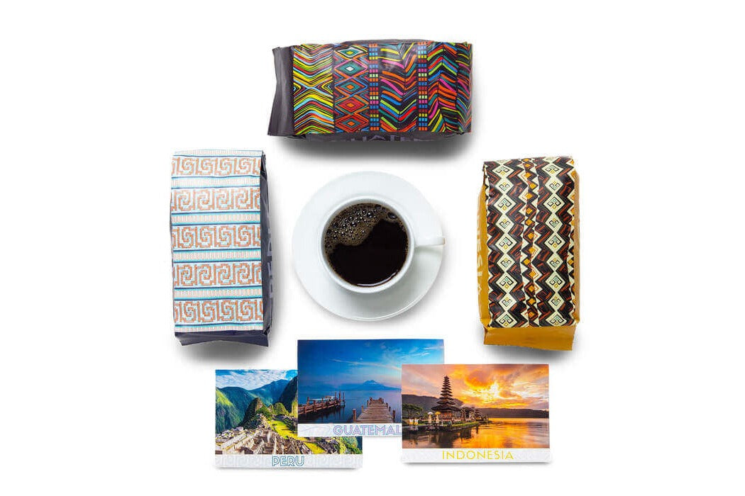 Three Atlas Coffee Club coffee bags in bold patterned packaging around a cup of black coffee, with postcards from Peru, Guatemala, and Indonesia