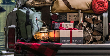 Three Bespoke Post branded boxes, two tied with red ribbon, in the back of a packed station wagon next to a lit lantern, a canvas duffel, and stacked firewood