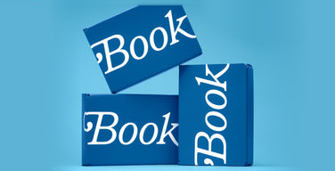 Three Book of the Month subscription boxes in the brand’s blue, each printed with the word Book in white serif type, stacked on a light blue background