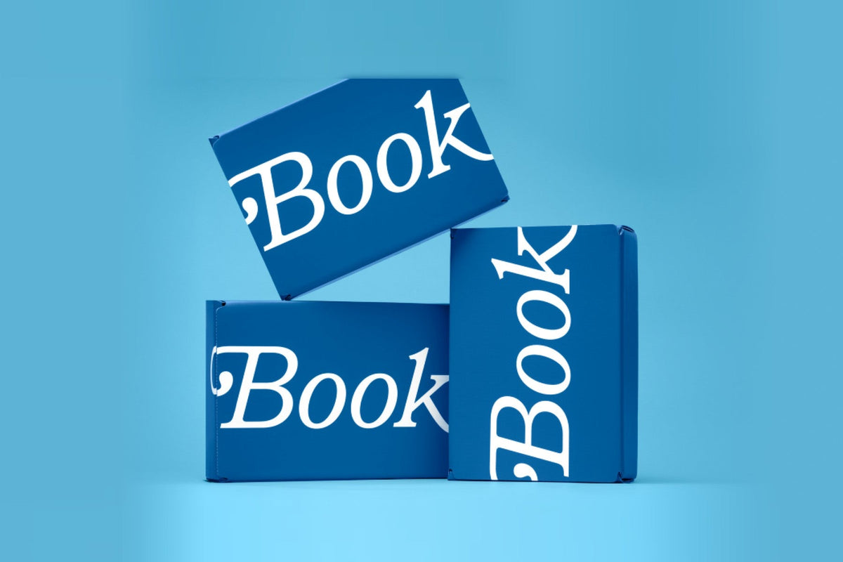 Three Book of the Month subscription boxes in the brand’s blue, each printed with the word Book in white serif type, stacked on a light blue background