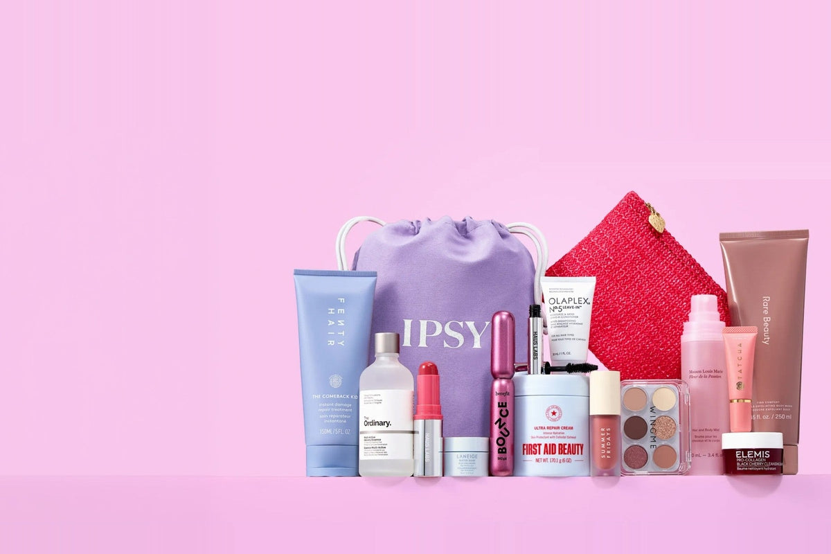 IPSY’s purple drawstring bag with the IPSY wordmark, surrounded by full-size beauty products including a Fenty Hair tube, The Ordinary serum, a Benefit mascara, First Aid Beauty cream, and a Wingme eyeshadow palette, on a pink background