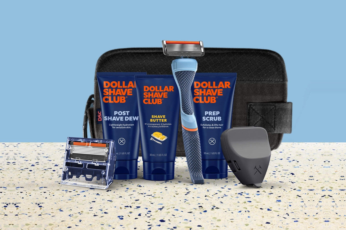 Dollar Shave Club's Fully Loaded Starter Set: Post Shave Dew, Shave Butter, and Prep Scrub tubes with the Dollar Shave Club logo, a blue handle with a six-blade cartridge, a cartridge refill, and a black travel case