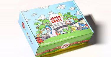 A Japan Crate box printed with the red Japan Crate logo, cartoon clouds, pine trees, a lucky cat, and a dragon, labeled as an assortment of Japanese candies and snacks plus a drink