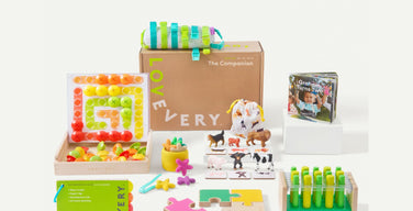 A Lovevery Play Kit box printed with the Lovevery logo and The Companion, surrounded by the kit’s wooden puzzle, peg board, animal figures, board book, and green Play Guide