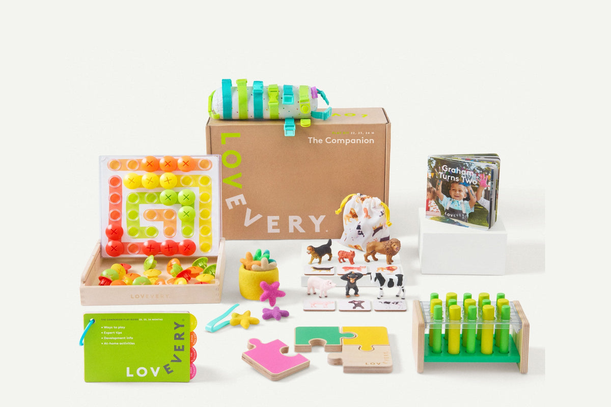 A Lovevery Play Kit box printed with the Lovevery logo and The Companion, surrounded by the kit’s wooden puzzle, peg board, animal figures, board book, and green Play Guide
