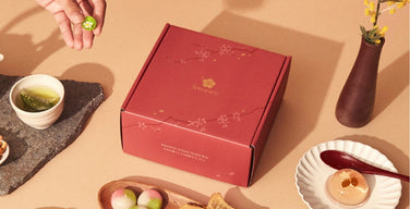 A red Sakuraco box printed with the Sakuraco logo and cherry blossoms, set among green tea, three-color mochi, taiyaki, and a clear jelly sweet
