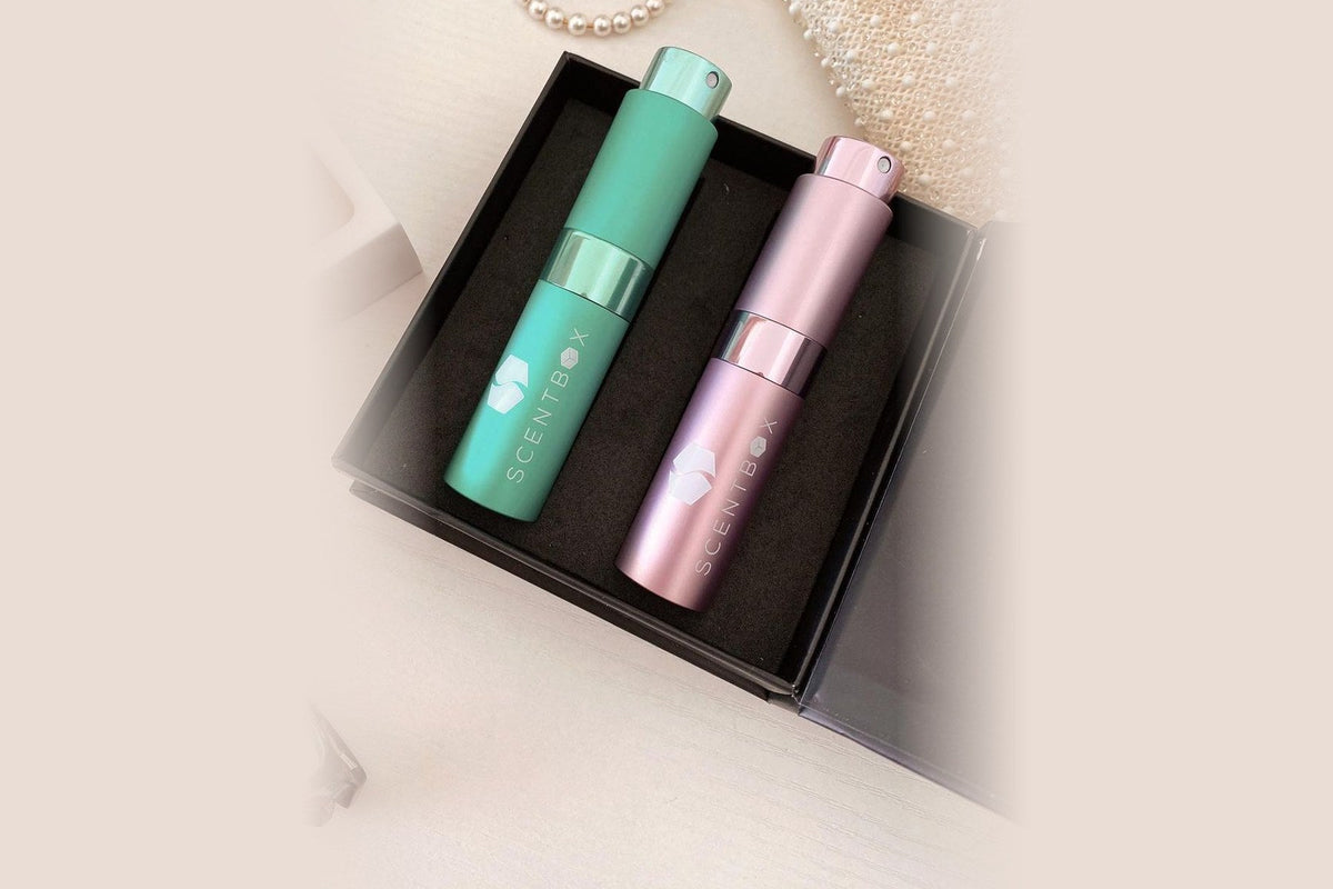 Two ScentBox perfume atomizers, one teal and one pink, each printed with the ScentBox logo, in an open black keepsake box on a cream tabletop