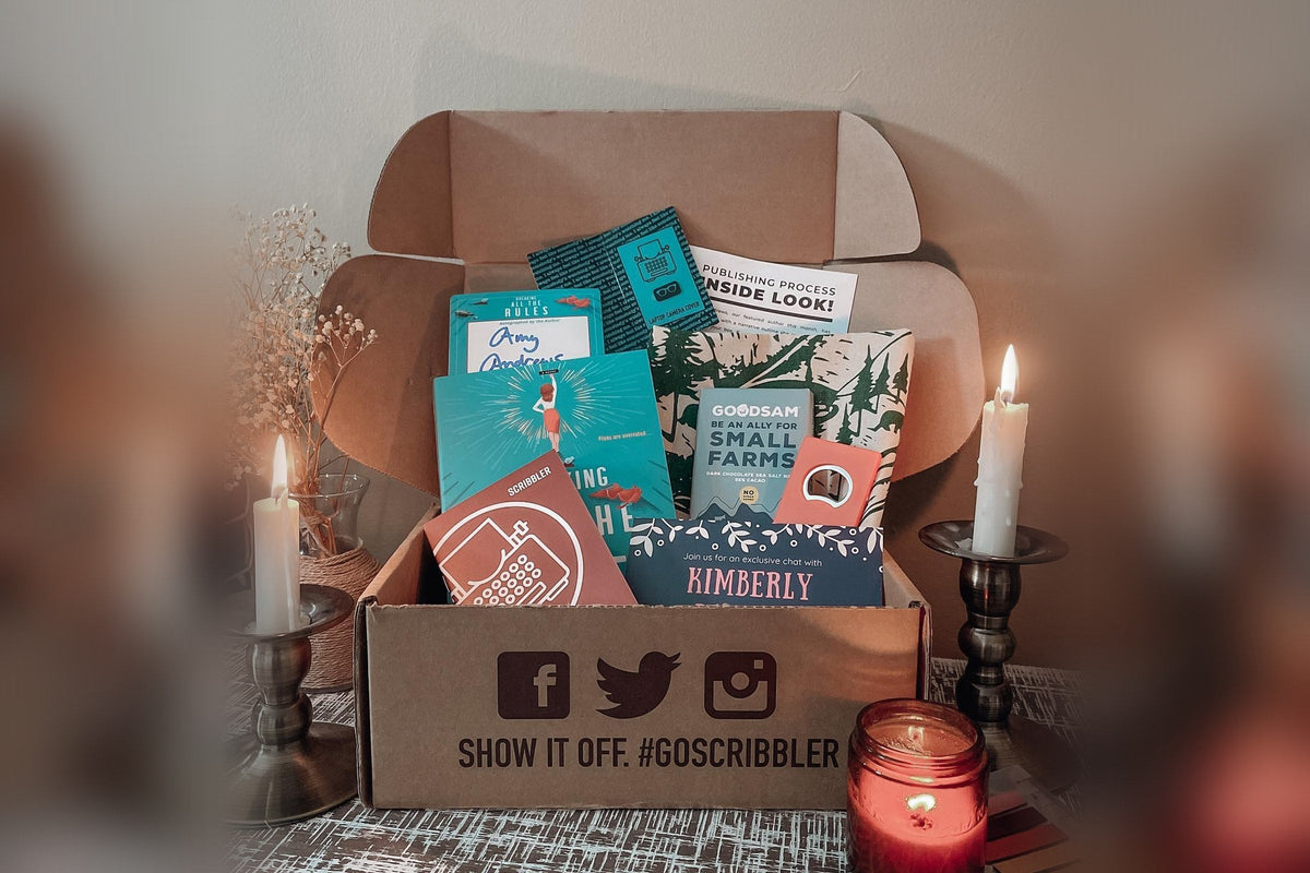 An open Scribbler box printed with “Show it off. #goscribbler,” holding a signed novel, a Scribbler writing passport with a typewriter logo, a publishing process card, and an author chat invite, between two lit candles