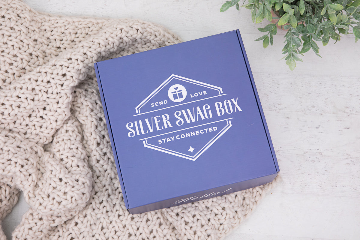 A blue Silver Swag Box with its lid on, printed with its Send Love, Stay Connected logo, resting on a cream knit blanket beside a small potted plant