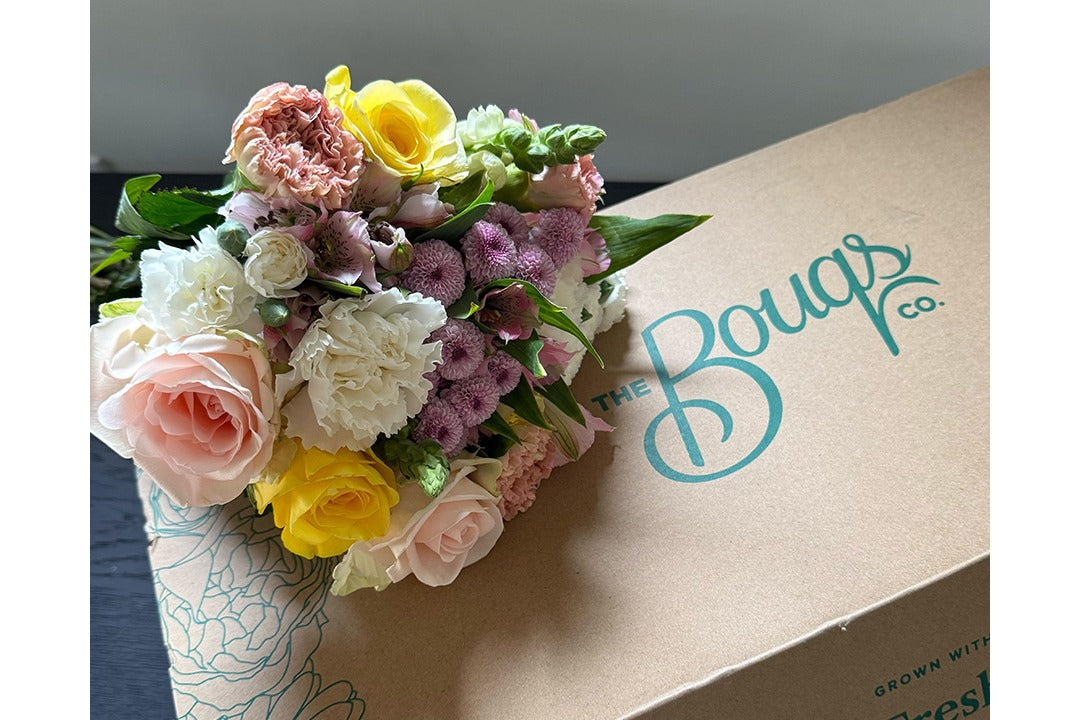 A pastel bouquet of roses, carnations, and button mums resting on a kraft delivery box printed with The Bouqs Co. logo in teal