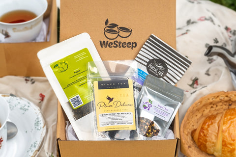 WeSteep Monthly Tea Box on a picnic table surrounded by cups of tea and pastries. Loose leaf tea pouches are featured inside the box.