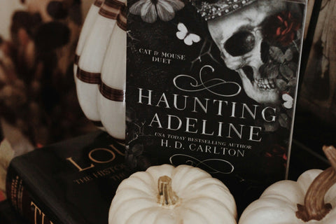 High quality image of horror books called haunting adeline from h d carlton stacked on each other with horror items around it.