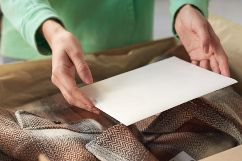 Woman's hands holding a blank card over a box filled with clothes. Woman is wearing a green shirt. Box has a brown blazer inside.