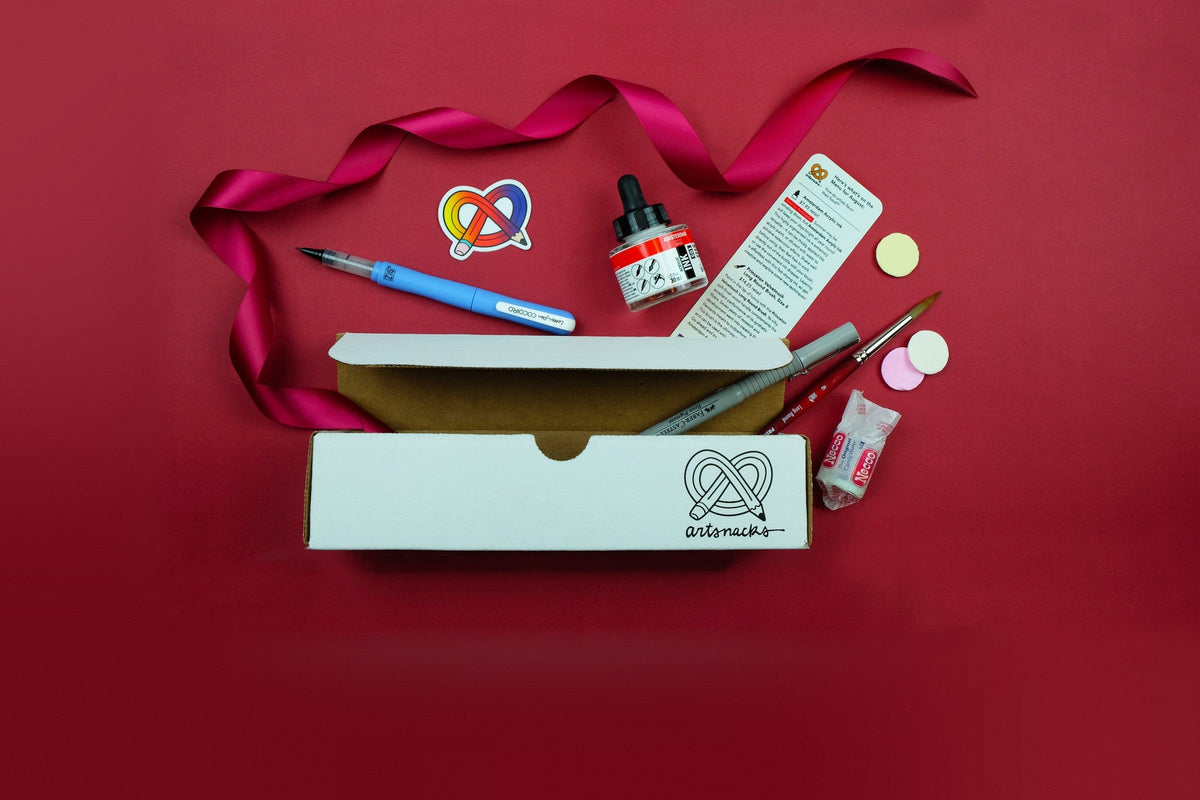 An open white ArtSnacks box printed with the pretzel-and-pencil artsnacks logo, with the August menu card, Amsterdam acrylic ink, a Zig letter pen, a Faber-Castell pen, a Princeton brush, a sticker, and Necco wafers on red paper