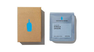 A Blue Bottle Coffee shipping box with the blue bottle logo beside a 12-ounce bag of Signature Single Origin whole bean coffee