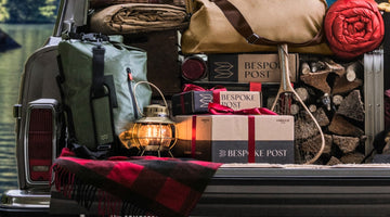 Three Bespoke Post branded boxes, two tied with red ribbon, in the back of a packed station wagon next to a lit lantern, a canvas duffel, and stacked firewood