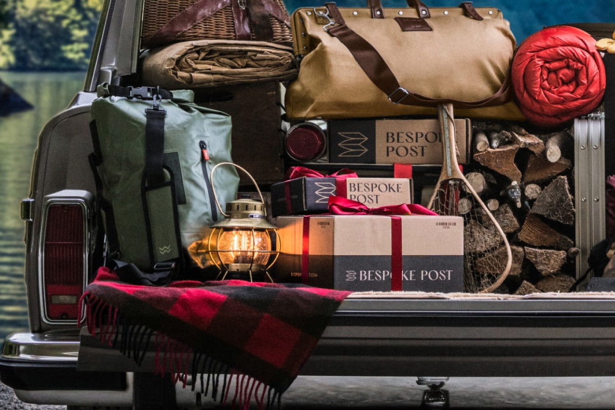 Three Bespoke Post branded boxes, two tied with red ribbon, in the back of a packed station wagon next to a lit lantern, a canvas duffel, and stacked firewood