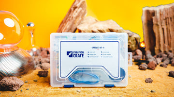 A clear Creation Crate project case on sand in front of a yellow backdrop, its label naming the Creation Crate 6 Project Kit and listing a mood lamp, memory game, distance detector, LED dice game, optical theremin, and 2-player reflex game