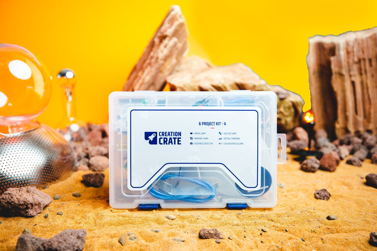 A clear Creation Crate project case on sand in front of a yellow backdrop, its label naming the Creation Crate 6 Project Kit and listing a mood lamp, memory game, distance detector, LED dice game, optical theremin, and 2-player reflex game