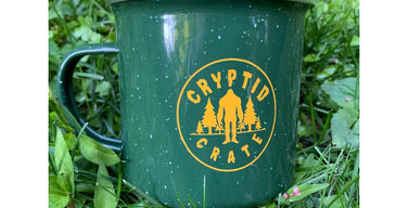 A green speckled enamel camp mug printed with the round yellow Cryptid Crate logo, a Bigfoot silhouette between pine trees, sitting in green grass