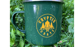 A green speckled enamel camp mug printed with the round yellow Cryptid Crate logo, a Bigfoot silhouette between pine trees, sitting in green grass