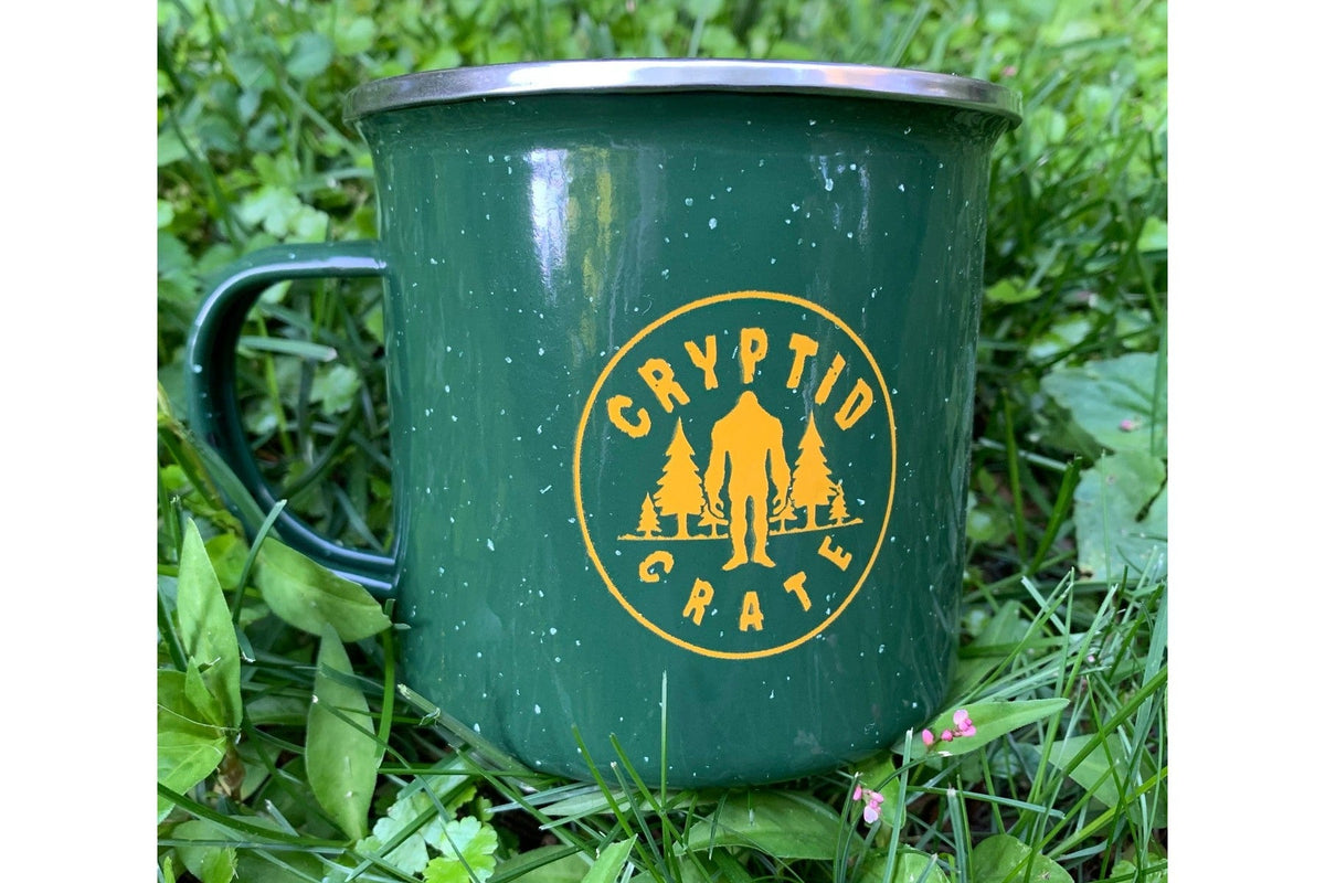 A green speckled enamel camp mug printed with the round yellow Cryptid Crate logo, a Bigfoot silhouette between pine trees, sitting in green grass