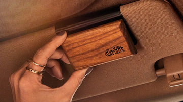 A hand clipping a Drift wood car freshener, engraved with the drift logo, onto a metal clip on a tan car sun visor