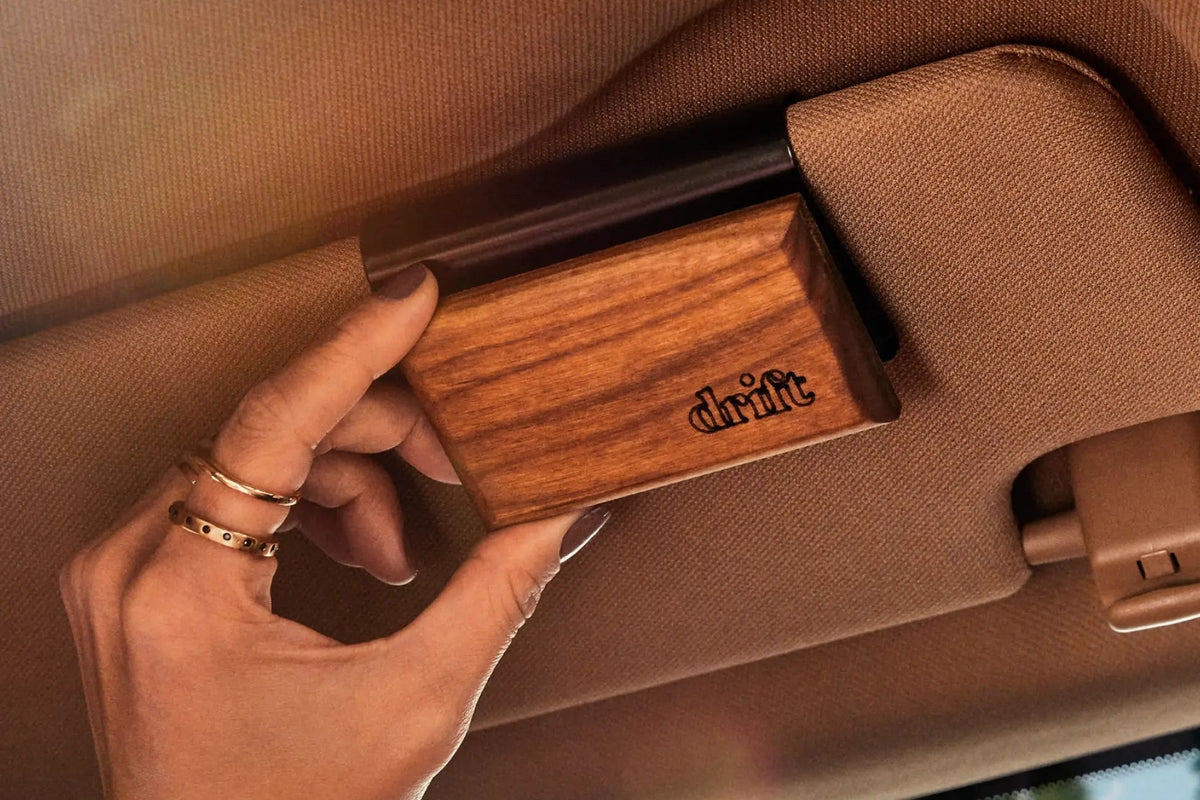 A hand clipping a Drift wood car freshener, engraved with the drift logo, onto a metal clip on a tan car sun visor