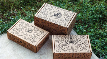 Three kraft FairyLoot subscription boxes printed in black with vines, moons, and a fairy emblem, the FairyLoot name on the side of the center box, set on stone beside grass