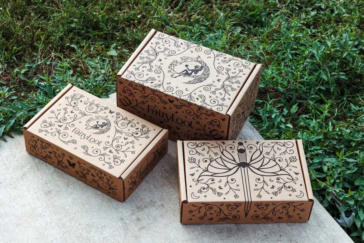Three kraft FairyLoot subscription boxes printed in black with vines, moons, and a fairy emblem, the FairyLoot name on the side of the center box, set on stone beside grass