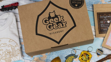 A kraft GeekGear Wizardry box with the Geek Gear cauldron logo and a Handmade With Love seal, beside a Hogwarts T-shirt, a toy Hogwarts Express, keyrings, a golden egg, chocolate frog tea, and a wand on a pale blue table