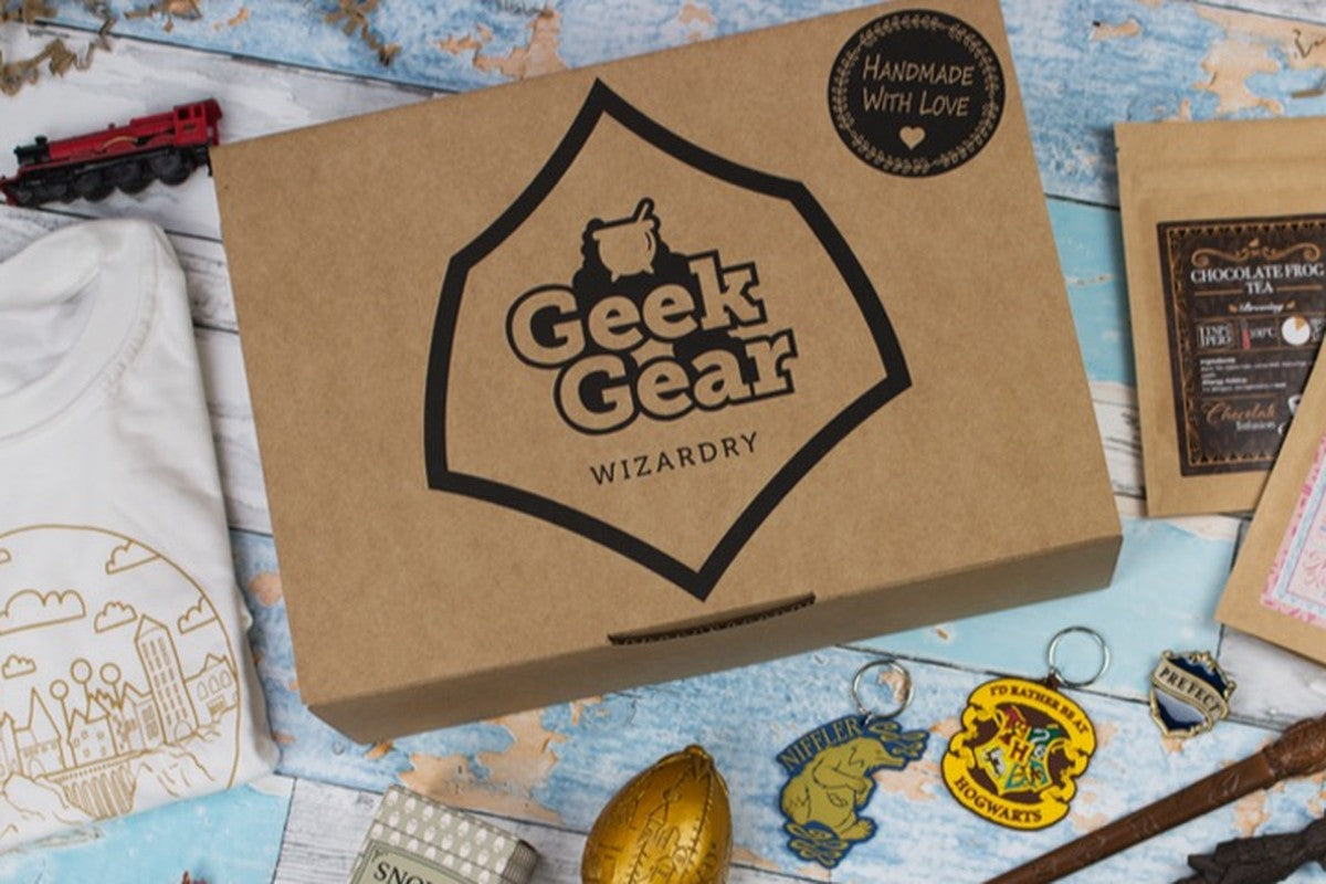 A kraft GeekGear Wizardry box with the Geek Gear cauldron logo and a Handmade With Love seal, beside a Hogwarts T-shirt, a toy Hogwarts Express, keyrings, a golden egg, chocolate frog tea, and a wand on a pale blue table