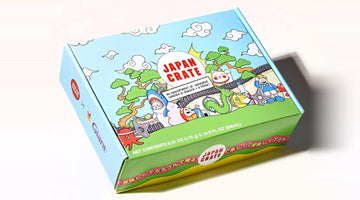A Japan Crate box printed with the red Japan Crate logo, cartoon clouds, pine trees, a lucky cat, and a dragon, labeled as an assortment of Japanese candies and snacks plus a drink