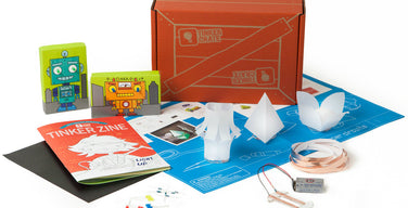 KiwiCo Tinker Crate box with its Tinker Zine booklet and a paper circuits kit: LEDs, copper tape, a 9-volt battery, an instruction sheet, and three folded paper lamp shades.