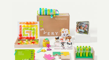 A Lovevery Play Kit box printed with the Lovevery logo and The Companion, surrounded by the kit’s wooden puzzle, peg board, animal figures, board book, and green Play Guide