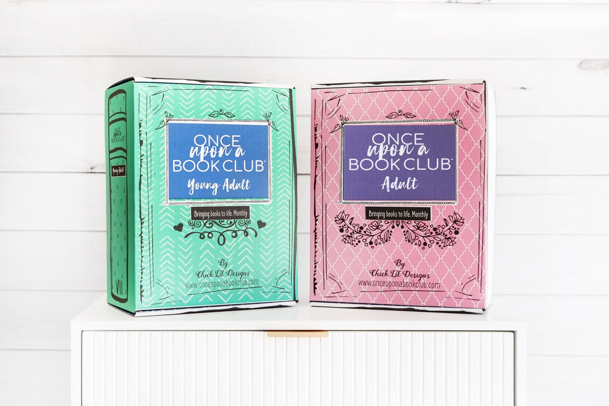 Two Once Upon a Book Club book-shaped boxes on a white dresser, a teal Young Adult box and a pink Adult box, each with the Once Upon a Book Club logo