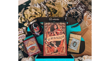 An open OwlCrate box with the owlcrate owl logo on its lid and “Curated for readers by readers” on the teal front, holding the hardcover All We Hunger For and themed bookish goodies on a fantasy map
