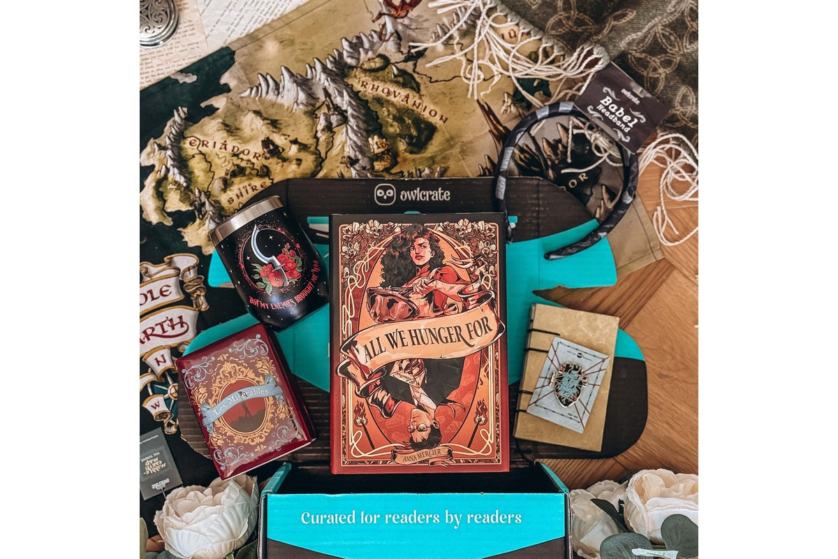 An open OwlCrate box with the owlcrate owl logo on its lid and “Curated for readers by readers” on the teal front, holding the hardcover All We Hunger For and themed bookish goodies on a fantasy map