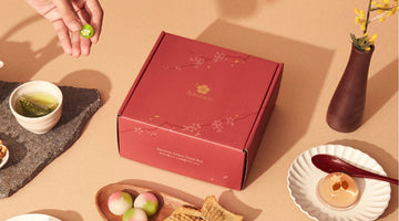 A red Sakuraco box printed with the Sakuraco logo and cherry blossoms, set among green tea, three-color mochi, taiyaki, and a clear jelly sweet
