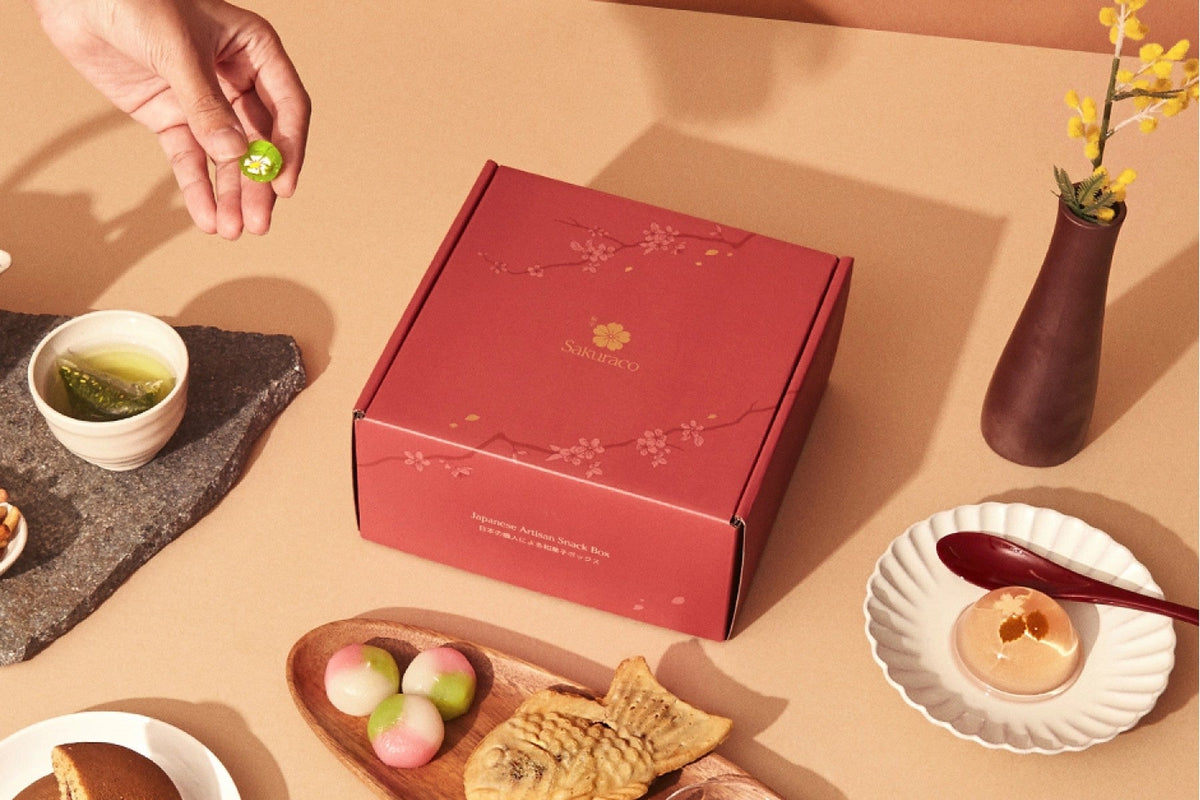 A red Sakuraco box printed with the Sakuraco logo and cherry blossoms, set among green tea, three-color mochi, taiyaki, and a clear jelly sweet