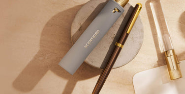 A gray Scentbird travel spray case with the gold Scentbird logo, resting on a stone dish beside a brown pen and a glass vial with a gold cap