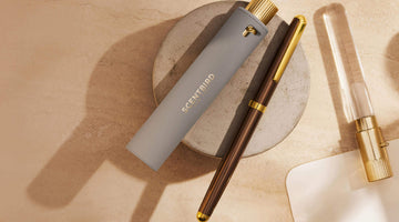 A gray Scentbird travel spray case with the gold Scentbird logo, resting on a stone dish beside a brown pen and a glass vial with a gold cap
