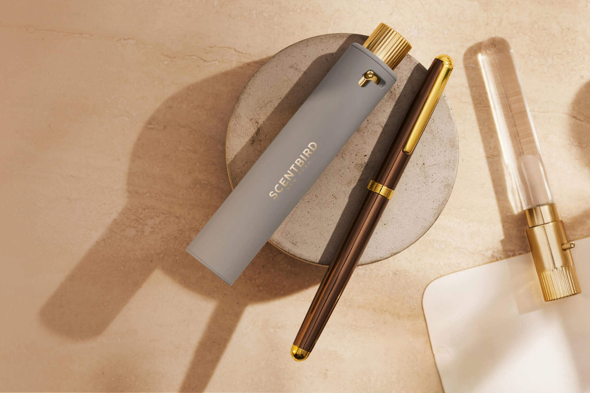 A gray Scentbird travel spray case with the gold Scentbird logo, resting on a stone dish beside a brown pen and a glass vial with a gold cap