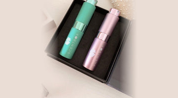 Two ScentBox perfume atomizers, one teal and one pink, each printed with the ScentBox logo, in an open black keepsake box on a cream tabletop