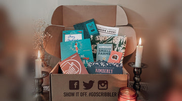 An open Scribbler box printed with “Show it off. #goscribbler,” holding a signed novel, a Scribbler writing passport with a typewriter logo, a publishing process card, and an author chat invite, between two lit candles