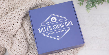 A blue Silver Swag Box with its lid on, printed with its Send Love, Stay Connected logo, resting on a cream knit blanket beside a small potted plant