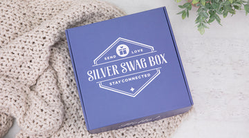 A blue Silver Swag Box with its lid on, printed with its Send Love, Stay Connected logo, resting on a cream knit blanket beside a small potted plant