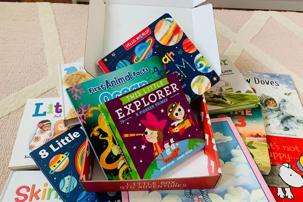 An open red-and-white box printed “Little box, big adventures” on its front edge, holding board books such as This Little Explorer and First Animal Facts: Ocean, with more children’s books spread on a rug around it.