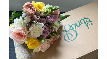 A pastel bouquet of roses, carnations, and button mums resting on a kraft delivery box printed with The Bouqs Co. logo in teal