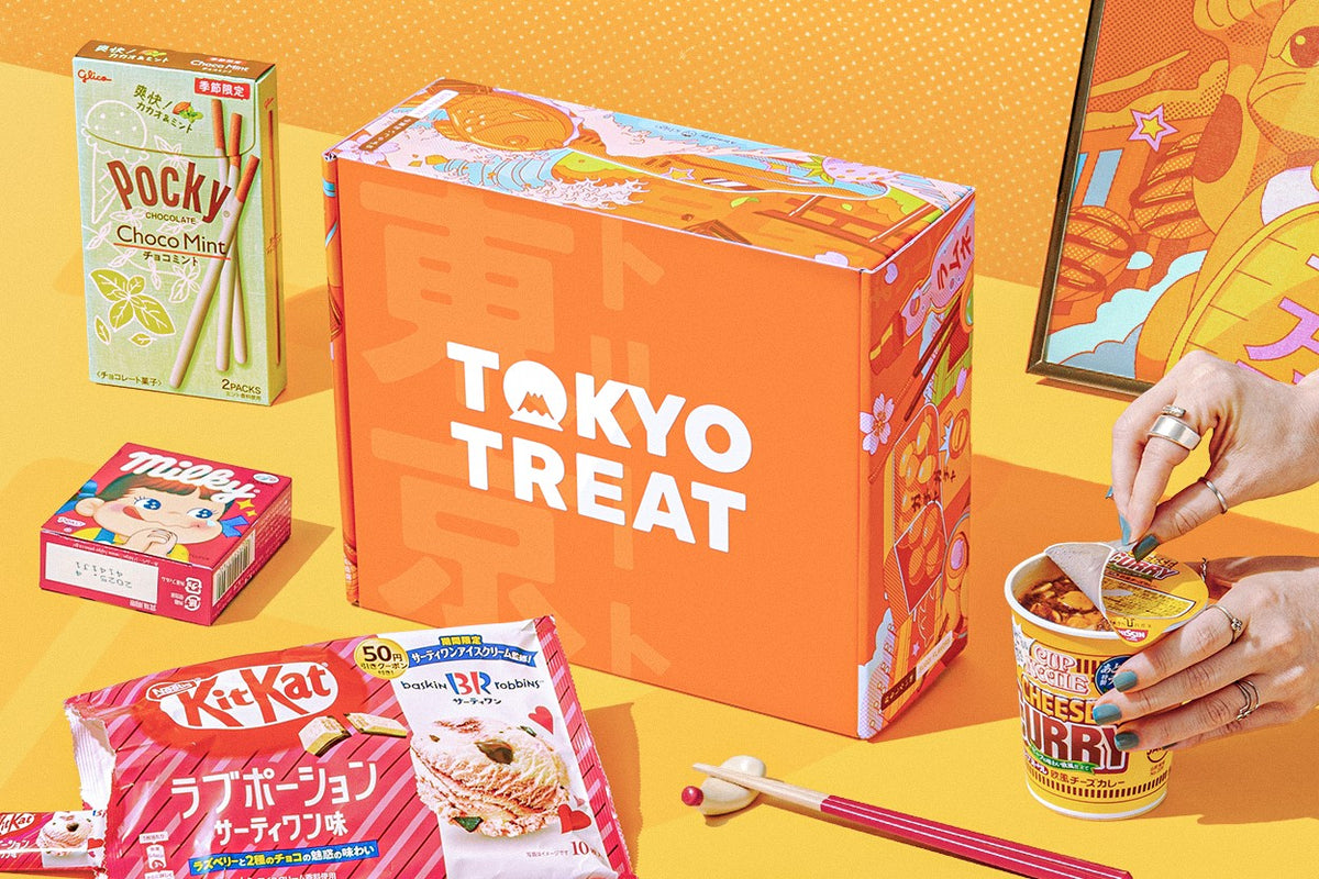 An orange TokyoTreat box printed with the TokyoTreat logo, surrounded by Japanese snacks including Choco Mint Pocky, a Baskin-Robbins Kit Kat, Milky candy, and Cup Noodle Cheese Curry